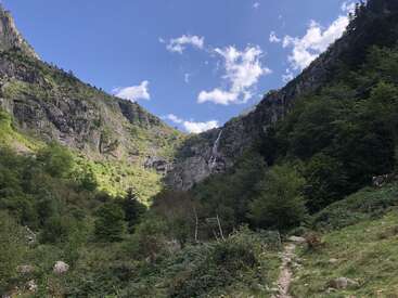 This image shows a scenic mountain valley with rocky cliffs, green trees, and a hidden waterfall. A narrow dirt path winds through the lush landscape under a blue sky.
