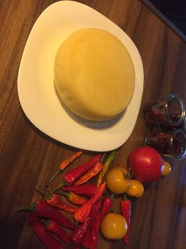 A round, yellow cheese wheel sits on a white plate. Colorful chili peppers, yellow and red tomatoes, and two jars of pickled vegetables surround it.