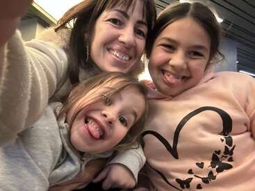 Uma selfie alegre de três pessoas: um adulto e duas crianças. Elas estão sorrindo de forma divertida, mostrando a língua, e parecem estar curtindo um momento divertido juntas.