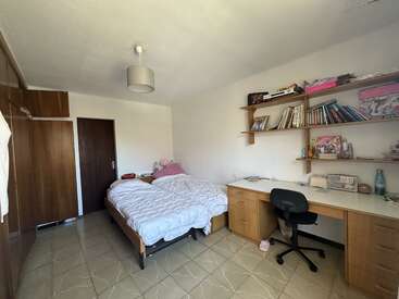 Um quarto arrumado com uma cama de solteiro, roupa de cama branca, móveis de madeira, estantes com livros, uma mesa com uma cadeira, piso de azulejo e pouca decoração.