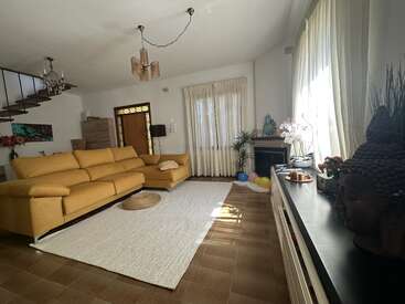 Uma aconchegante sala de estar tem um sofá modular amarelo mostarda, um tapete branco, janelas amplas com cortinas, plantas de interior, uma cabeça de Buda decorativa e balões coloridos e divertidos perto da lareira.