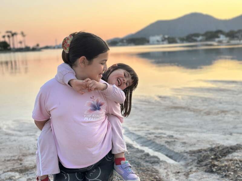 Duas crianças sorridentes brincam à beira de um lago tranquilo ao pôr do sol. A menina mais velha carrega a mais nova nas costas, e as duas riem, com montanhas e palmeiras ao fundo.