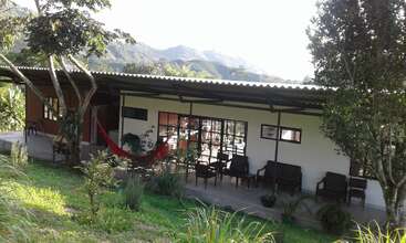A modern, single-story house with a large porch, comfortable outdoor seating, a red hammock, surrounded by lush greenery and trees, set against scenic mountain views.