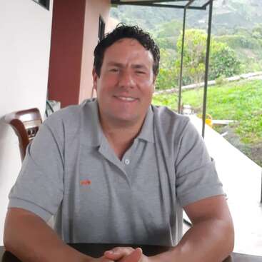 A smiling man with short curly hair sits at a table outdoors, wearing a grey polo shirt. Lush greenery and a building can be seen behind him.