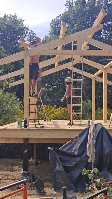 Two people are building a wooden structure, standing on ladders and a platform, surrounded by trees and construction tools, working under a sunny sky outdoors.