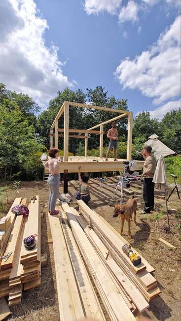 Four people and a dog are building a wooden structure outdoors. Lumber, tools, and supplies are scattered around. The sky is blue with scattered white clouds.
