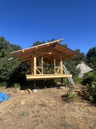 A small wooden structure under construction stands elevated on pillars in a clearing, surrounded by green trees and clear blue sky, with building materials nearby.