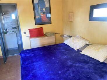 Este acogedor dormitorio cuenta con una cama grande con una manta de un azul intenso, paredes amarillas, obras de arte modernas, una ventana pequeña y un cuarto de baño contiguo con azulejos grises.