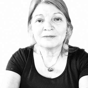 Un retrato en blanco y negro de una mujer de mediana edad con el pelo claro, que lleva una camiseta oscura, un collar con colgante y pendientes de aro, y que mira directamente a la cámara.