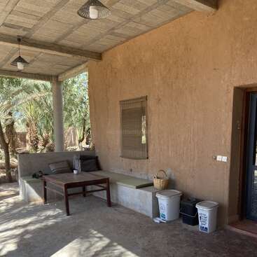 La imagen muestra un sereno espacio al aire libre con un banco, una mesa y papeleras, rodeado de palmeras y un muro rústico, que evoca una sensación de tranquilidad y belleza natural.