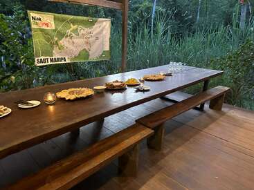 A long wooden table set outdoors displays plates of food, glassware, and a candle. A map labeled "Saut Maripa" hangs nearby. Lush greenery surrounds.
