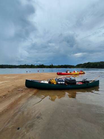 A green canoe loaded with gear rests on a sandy shore, with another boat nearby. Swimmers enjoy the water under a cloudy, dramatic sky. Peaceful nature.