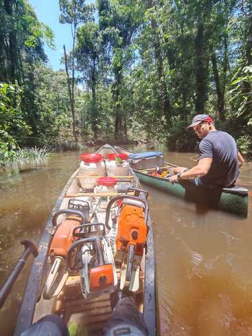 A man paddles a canoe through a jungle river, transporting chainsaws, fuel, and supplies. Sunlight filters through dense trees, creating a remote, adventurous scene.