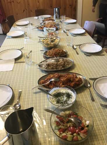 Une longue table est dressée pour huit personnes, avec divers plats, notamment du riz, de la salade et du yaourt, prêts à être dégustés en commun dans une salle confortable.