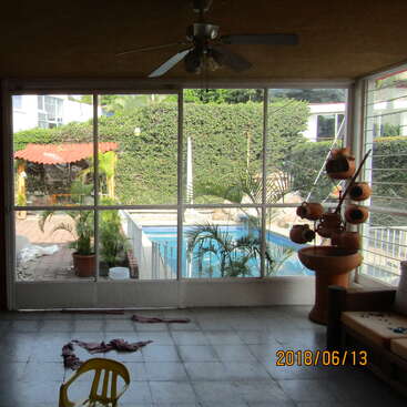 Das Bild zeigt einen Raum mit einem Tisch und Stühlen, in dem im Hintergrund ein Bücherregal steht. Der Raum ist gut beleuchtet, mit einem großen Fenster auf der linken Seite des Bildes.