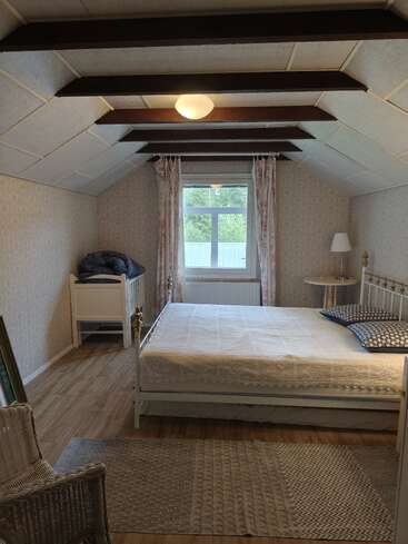 Cette chambre mansardée confortable comprend un lit double, un petit lit de repos, une chaise en osier, un tapis à motifs, des rideaux à fleurs, des poutres en bois apparentes et une lampe près d'une fenêtre lumineuse.
