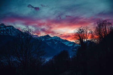 Un magnifique coucher de soleil illumine les montagnes enneigées et les silhouettes d'arbres, avec des teintes vibrantes de violet, de rose et d'orange peintes sur le ciel dramatique et nuageux. Une beauté tranquille.