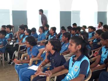 Un groupe d'écoliers en uniforme bleu est assis attentivement sur des chaises, écoutant et prenant des notes dans une salle de classe ou lors d'un événement éducatif, en présence d'enseignants.