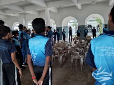 Un groupe d'étudiants en uniforme bleu se tient en demi-cercle à l'intérieur d'une salle avec des chaises en plastique, participant à une discussion ou à une activité en classe. Derrière, des fenêtres cintrées.