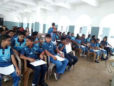 Un groupe d'étudiants portant des uniformes bleus sont assis sur des chaises en plastique à l'intérieur d'un hall spacieux et lumineux. Certains tiennent des documents, discutent et attendent un événement.