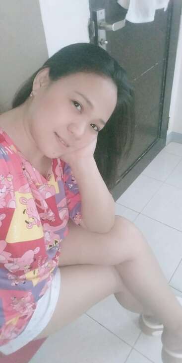 Une femme est assise à l'intérieur, la tête appuyée sur sa main, souriante. Elle porte une chemise colorée de la Panthère rose et un short blanc, avec une porte en arrière-plan.