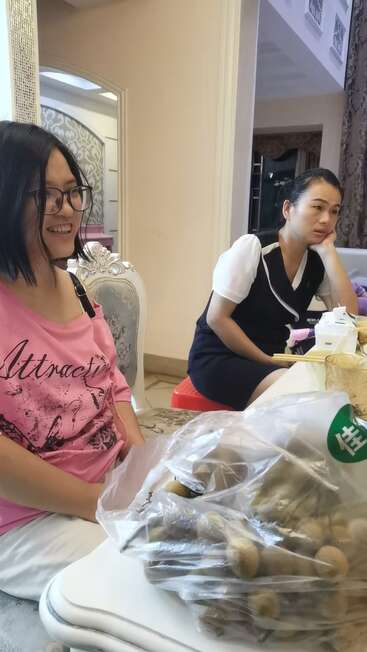 Deux femmes sont assises dans une pièce bien éclairée ; l'une sourit en portant une chemise rose, l'autre a l'air pensif. Un sac en plastique contenant des fruits longan est posé sur la table.