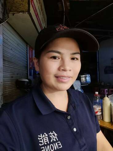 Une femme portant un polo bleu marine et une casquette noire se tient à l'intérieur, souriant légèrement à l'appareil photo. L'arrière-plan est composé de bouteilles, de récipients alimentaires et de volets de magasins fermés.