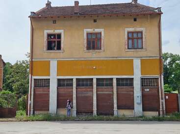 This image shows an old, yellowish two-story building with peeling paint. One person stands outside, while another looks out from an upstairs window. Bushes surround it.