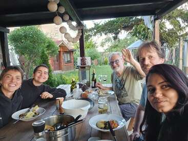 Five people are enjoying a meal together outdoors at a wooden table under a pergola. Everyone is smiling, and the atmosphere looks warm, relaxed, and friendly.