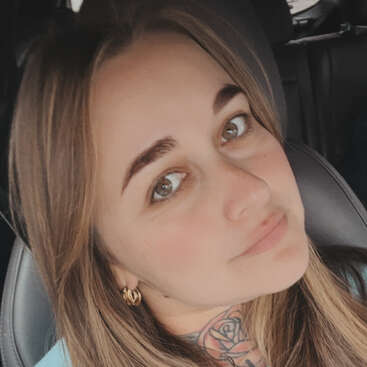 A woman with light brown hair and hazel eyes sits in a car seat, wearing gold hoop earrings. She has a rose tattoo on her neck.