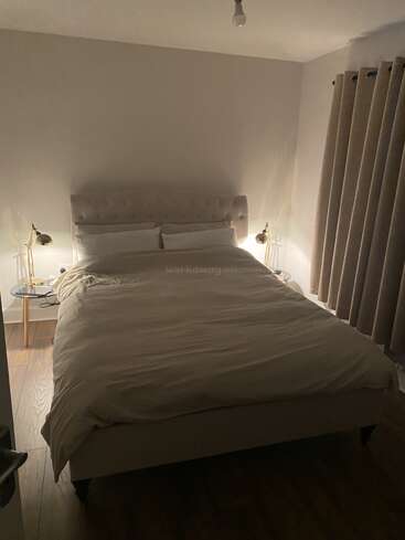 A cozy bedroom with a large bed, beige bedding, two bedside lamps, wooden floor, round side tables, thick curtains, and soft lighting creating a warm ambiance.