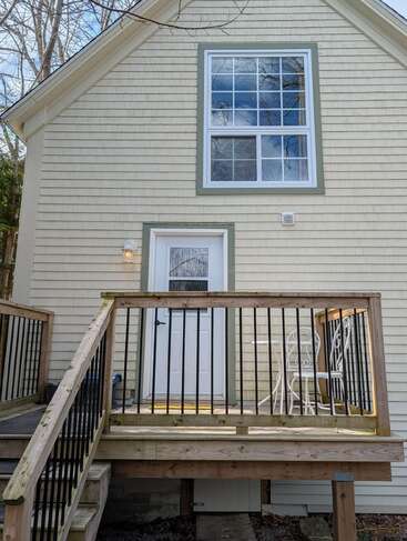 A small wooden porch with black railings leads to a white door. There's a white metal table and chair set. Beige house siding, large window above.