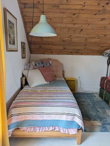A cozy attic bedroom with a single bed, colorful pillows, striped blanket, wood-paneled slanted ceiling, hanging light, framed pictures, rug, and stacked vintage suitcases.