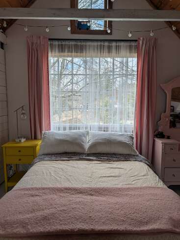 A cozy bedroom with a large window, pink curtains, pastel bedding, yellow nightstand, string lights, wooden beams, and a scenic view of trees outside.