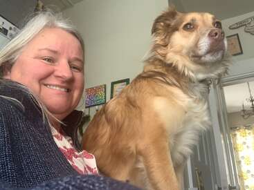 A smiling woman with gray hair takes a selfie indoors. A fluffy brown and white dog sits beside her, looking alert. Bright, cozy room background.