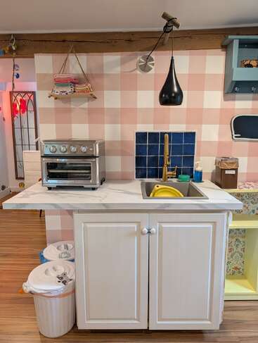 Esta acogedora cocina cuenta con un fregadero con mueble blanco, un horno tostador, cubos de basura, una lámpara colgante, una pared a cuadros, un salpicadero de azulejos azules y paños de cocina cuidadosamente doblados.
