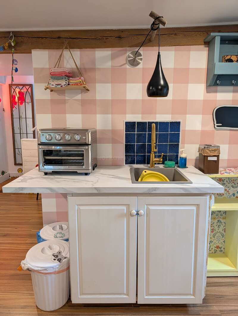 This cozy kitchen features a white cabinet sink, toaster oven, trash bins, hanging lamp, checkered wall, blue tile backsplash, and neatly folded kitchen towels.