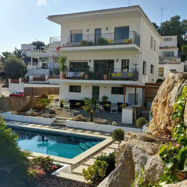 The image depicts a three-story white building with a balcony and a pool in the foreground, surrounded by lush greenery and a serene atmosphere.