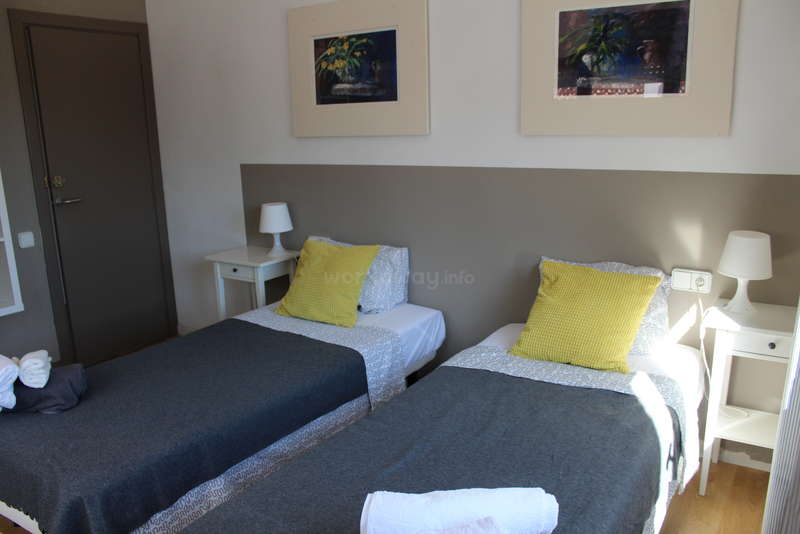 A imagem mostra um quarto com uma cama e uma mesa, com uma grande janela e uma porta, com uma parede branca e um piso cinza, proporcionando uma atmosfera limpa e simples.