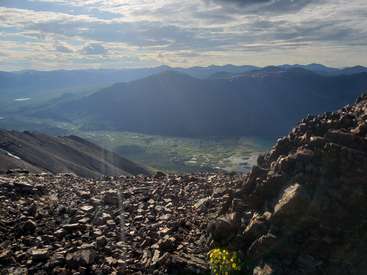 The image depicts a rocky mountainous landscape with a valley below, featuring a small patch of yellow flowers in the foreground and a cloudy sky above.