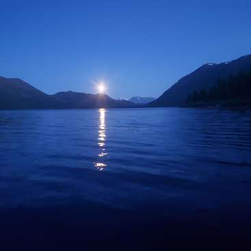 The image depicts a serene lake scene at dusk, with a sun setting behind mountains and its reflection visible on the water\'s surface.
