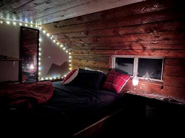 The image depicts a cozy bedroom with a bed, a window, and a mirror, illuminated by string lights and a lamp, creating a warm and inviting atmosphere.