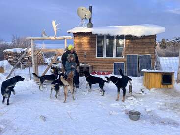 The image depicts a man and a child standing outside a small wooden cabin, surrounded by dogs, with a snowy landscape and a satellite dish on the roof.