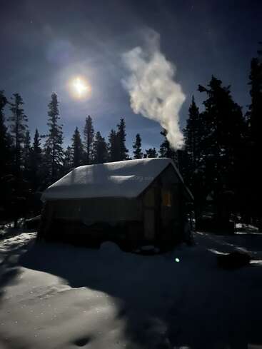 The image depicts a snow-covered cabin in the woods at night, with a bright moon shining above and smoke rising from the chimney.