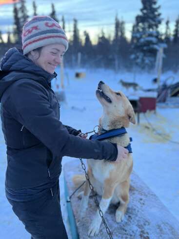 The image depicts a woman in winter attire petting a dog on a snowy surface, with trees and a sled in the background, conveying a sense of warmth and companionship.