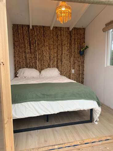 Das Bild zeigt ein gemütliches Schlafzimmer mit einem Bett, einem Kronleuchter und einer Bambuswand. Weiße Wände, ein Fenster und helle Holzböden vermitteln ein Gefühl von Wärme und Komfort.