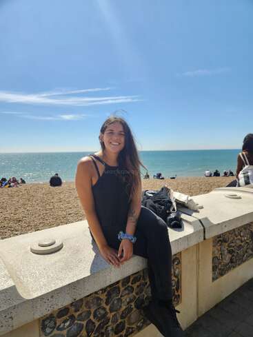 Das Bild zeigt eine Frau, die auf einer Steinmauer am Strand sitzt, mit einem ruhigen Blick auf das Meer und einem klaren blauen Himmel im Hintergrund.