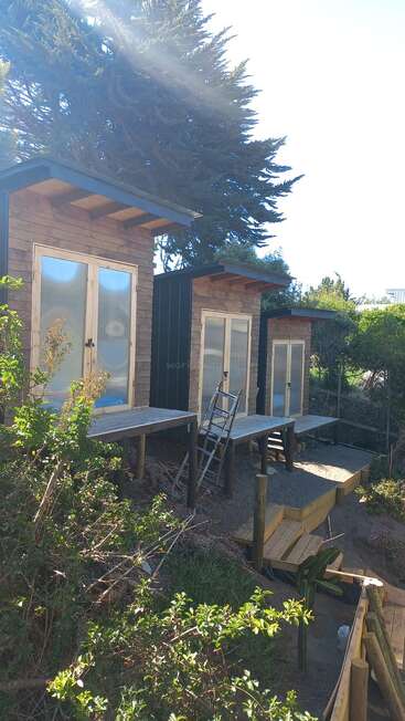 Das Bild zeigt ein kleines Holzhaus mit einer Terrasse, umgeben von Bäumen und Sträuchern, mit einer Leiter und einer Bank, vor einem hellen und sonnigen Himmel.