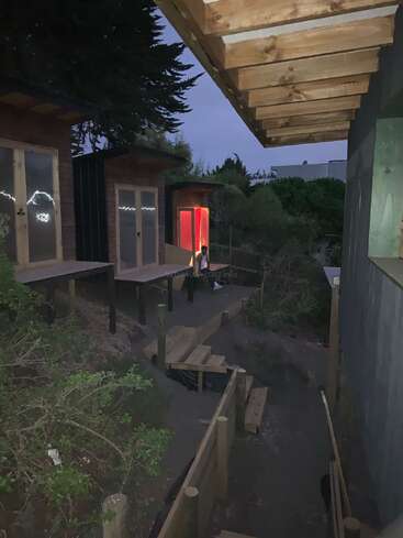 Das Bild zeigt eine ruhige Umgebung in der Abenddämmerung mit einem Haus, einer Terrasse, Holzbänken und einer Person, die auf dem Boden sitzt, umgeben von Bäumen und Büschen.