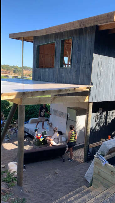 Das Bild zeigt eine hölzerne Konstruktion mit einem Deck, unter dem ein Kühlschrank und Menschen versammelt sind, während ein Hund in der Nähe auf dem Boden liegt.